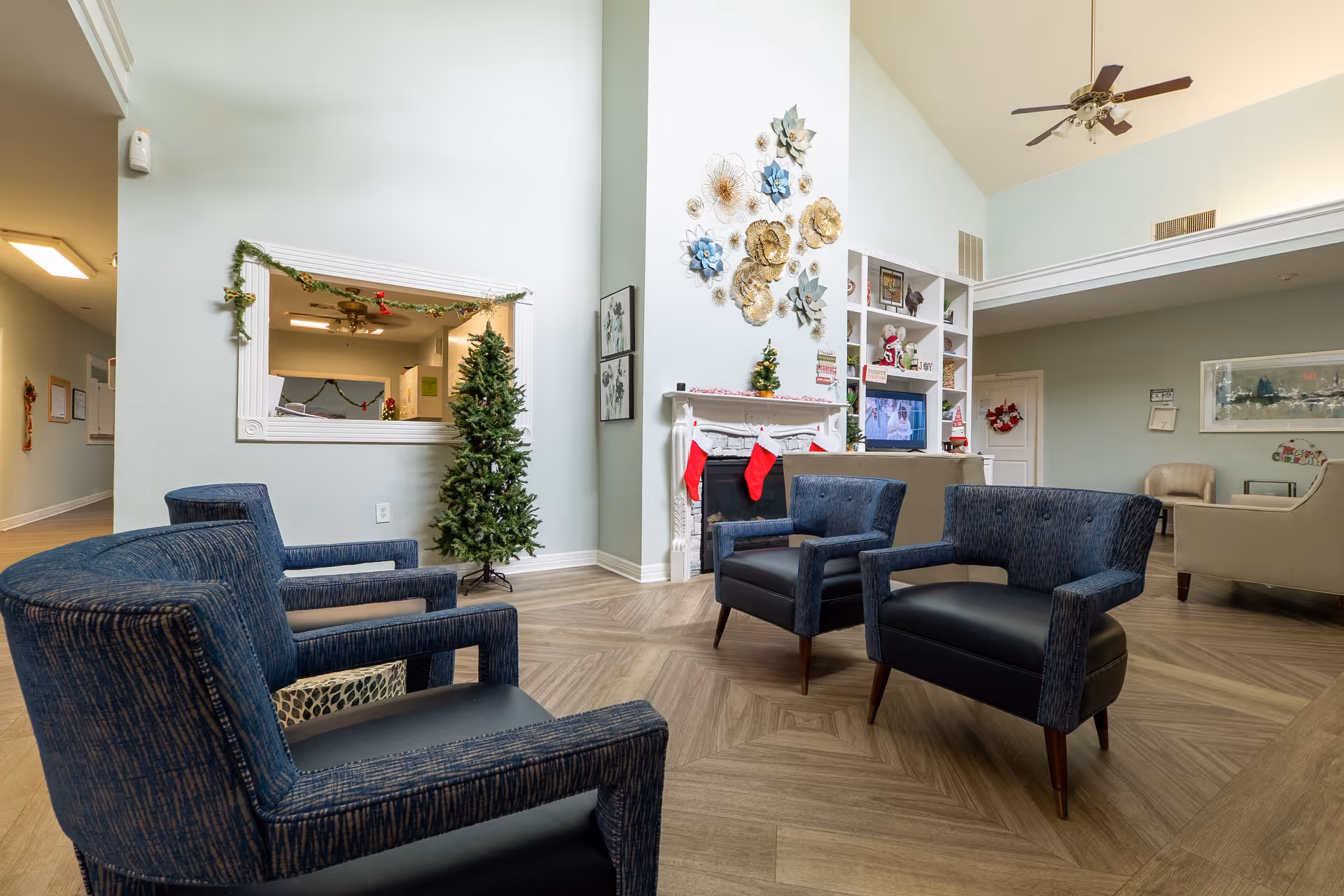 A senior living facility common area with blue upholstered chairs arranged around a white fireplace decorated with Christmas stockings. There is a Christmas tree next to the fireplace and festive garlands hanging around a window opening in the wall. The room has light-colored walls, a ceiling fan, and built-in shelves with holiday decorations and a television.