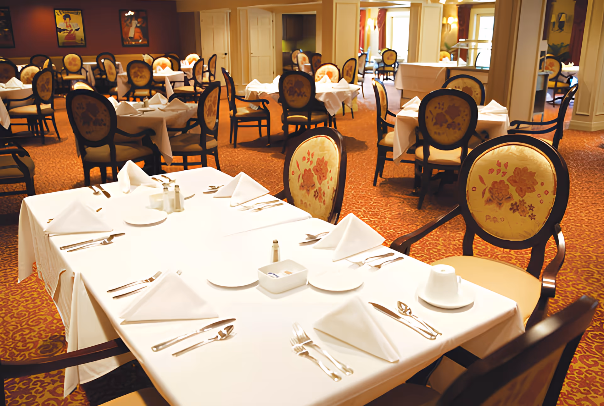 A formal dining room with multiple tables set with white tablecloths, folded napkins, and floral-upholstered chairs.