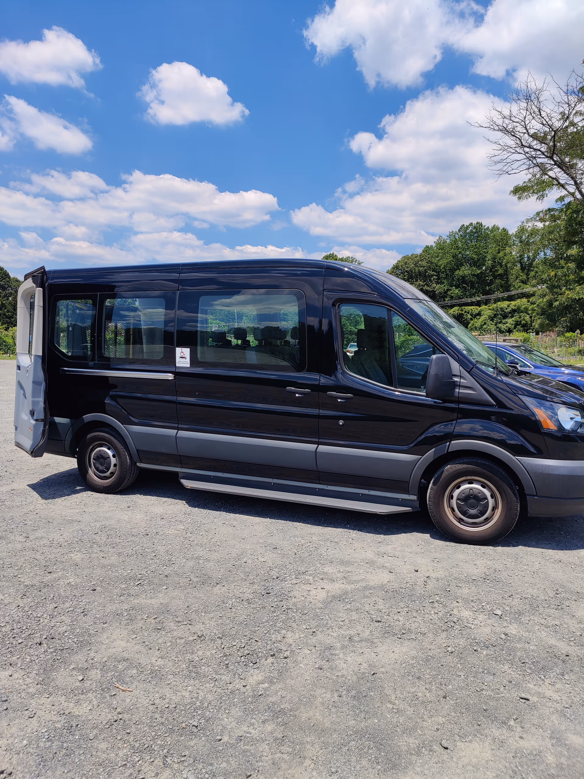 Black passenger van parked on a gravel lot, blue sky with clouds and trees in background.