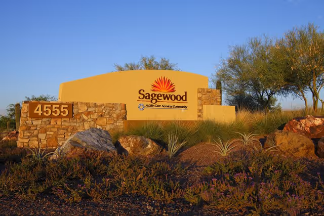 Stone and stucco sign for Sagewood ALife Care Services Community with the address number 4555, surrounded by desert landscaping including rocks, shrubs, and small trees under a clear blue sky.