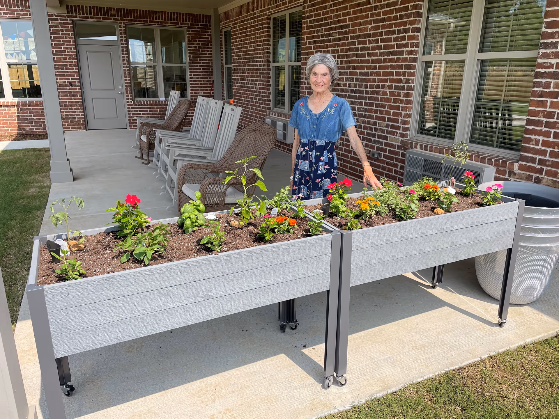 An elderly woman standing outdoors next to two raised garden beds filled with various flowering plants and greenery. Behind her is a covered patio area with several rocking chairs and a brick building with windows and a door.