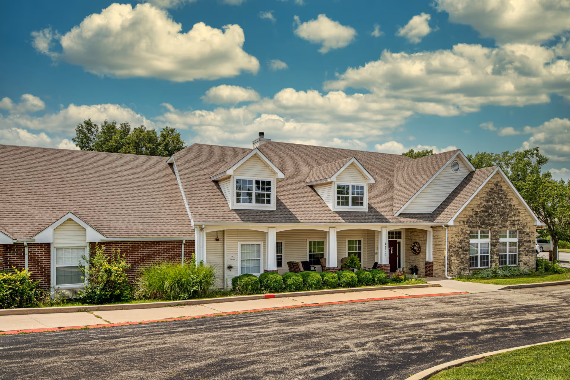 Front exterior of Creekside Village single-story residential building with a covered porch, dormer windows, and landscaped shrubs under a partly cloudy sky.