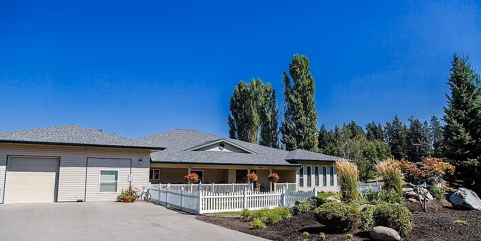 Single-story assisted living building with a white picket fence, landscaped beds and trees under a clear blue sky.
