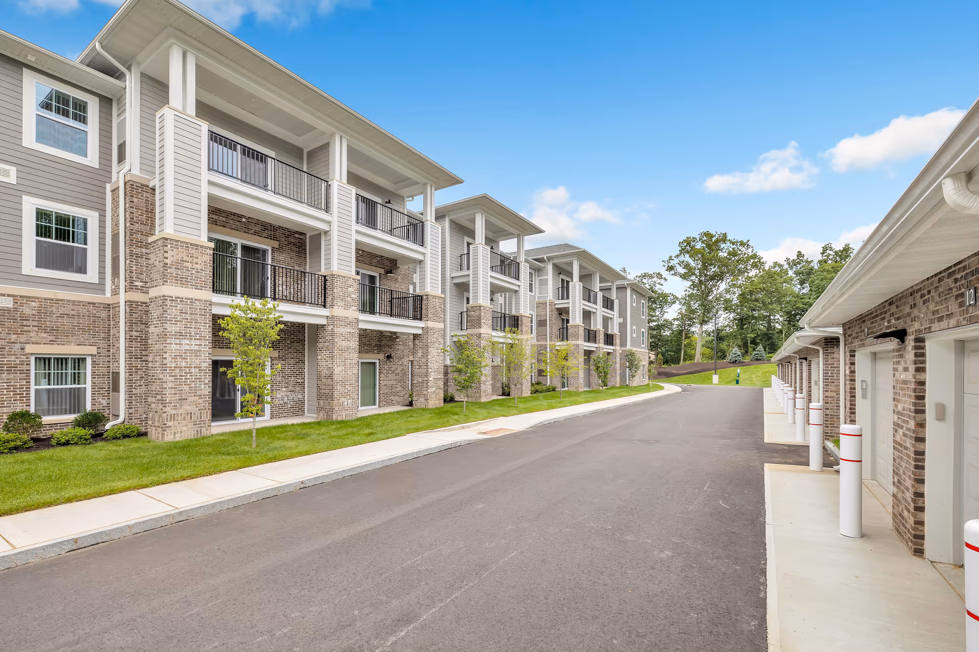 Three-story brick-and-siding senior living buildings with balconies, a paved driveway and garage bays under a clear blue sky.