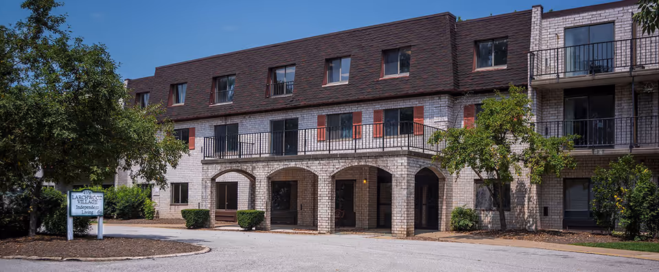 Front exterior of a three-story brick senior living building with arched entrance, balconies, trees, and a driveway.
