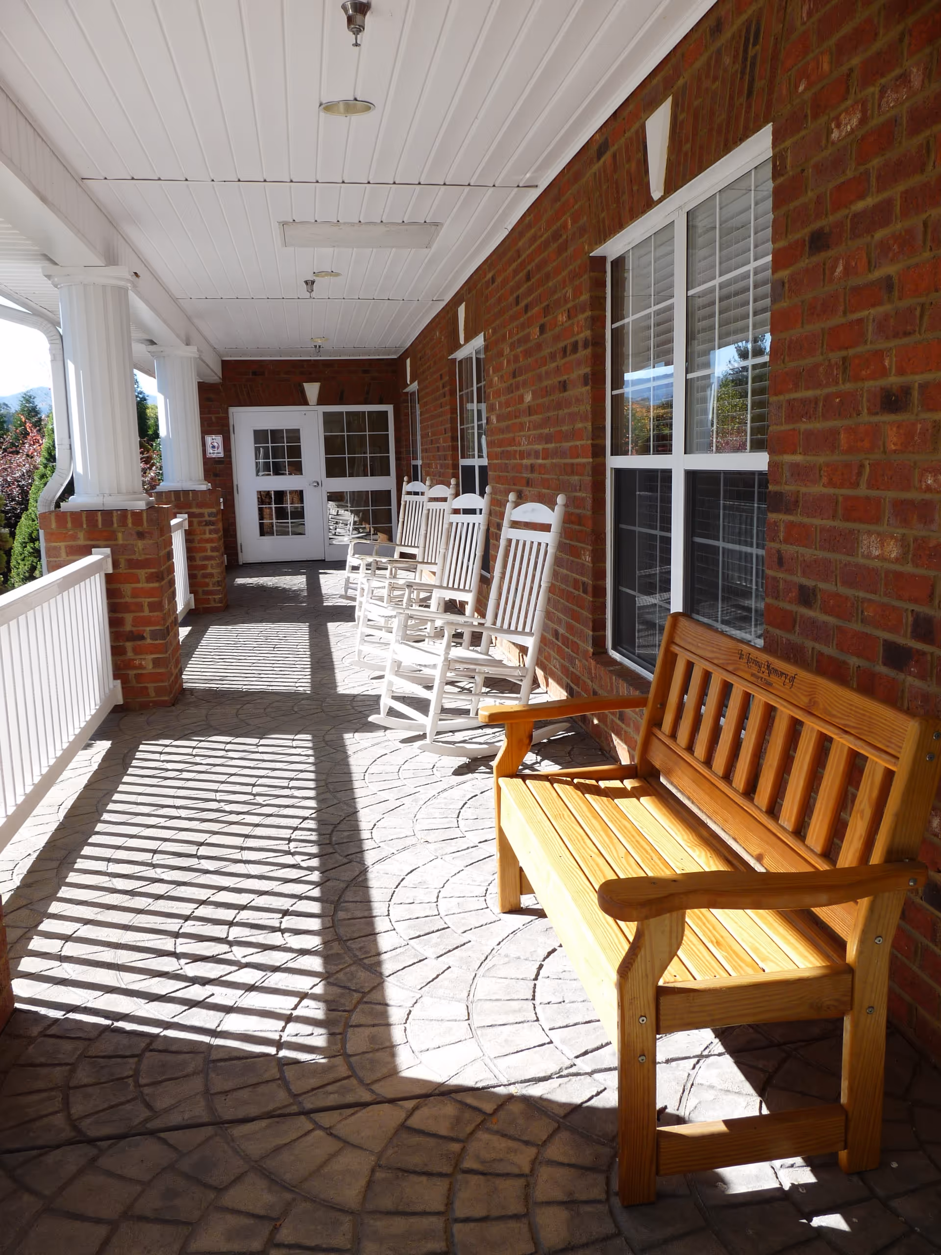 Brick exterior porch with white rocking chairs and a wooden bench beside large windows.