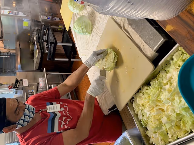 Staff member wearing mask and gloves chops cabbage on a cutting board in a busy kitchen.