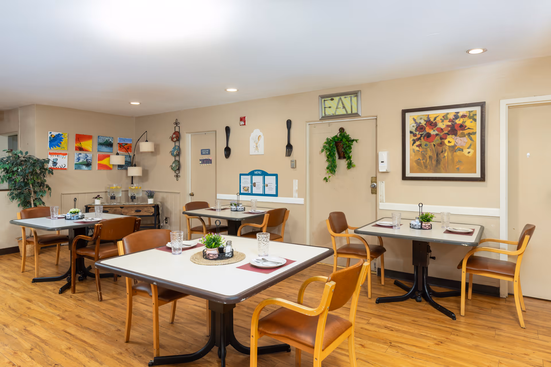A bright senior living dining room with multiple square tables, chairs, and wall art.