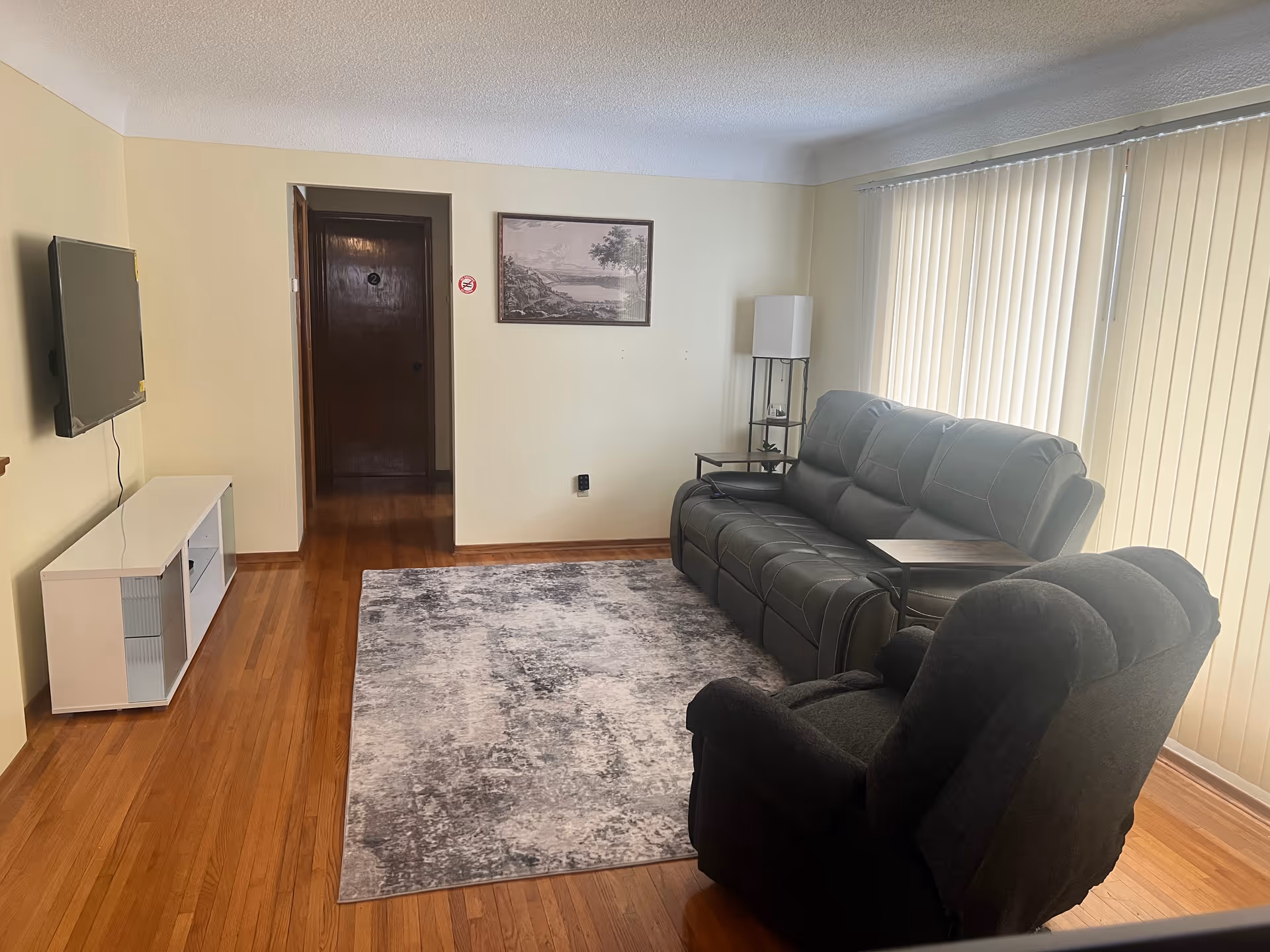 Cozy living room with black leather sofa and recliner, rug, TV, and vertical blinds.