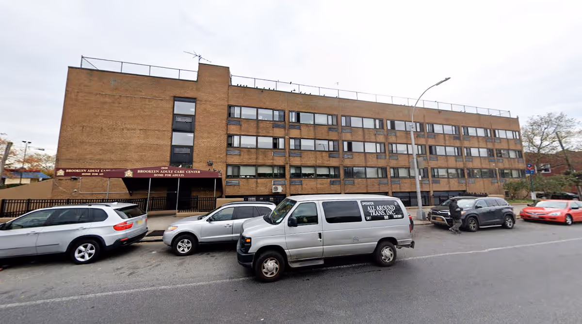 Front exterior of a multi-story brick building labeled Brooklyn Adult Care Center with cars parked along the street
