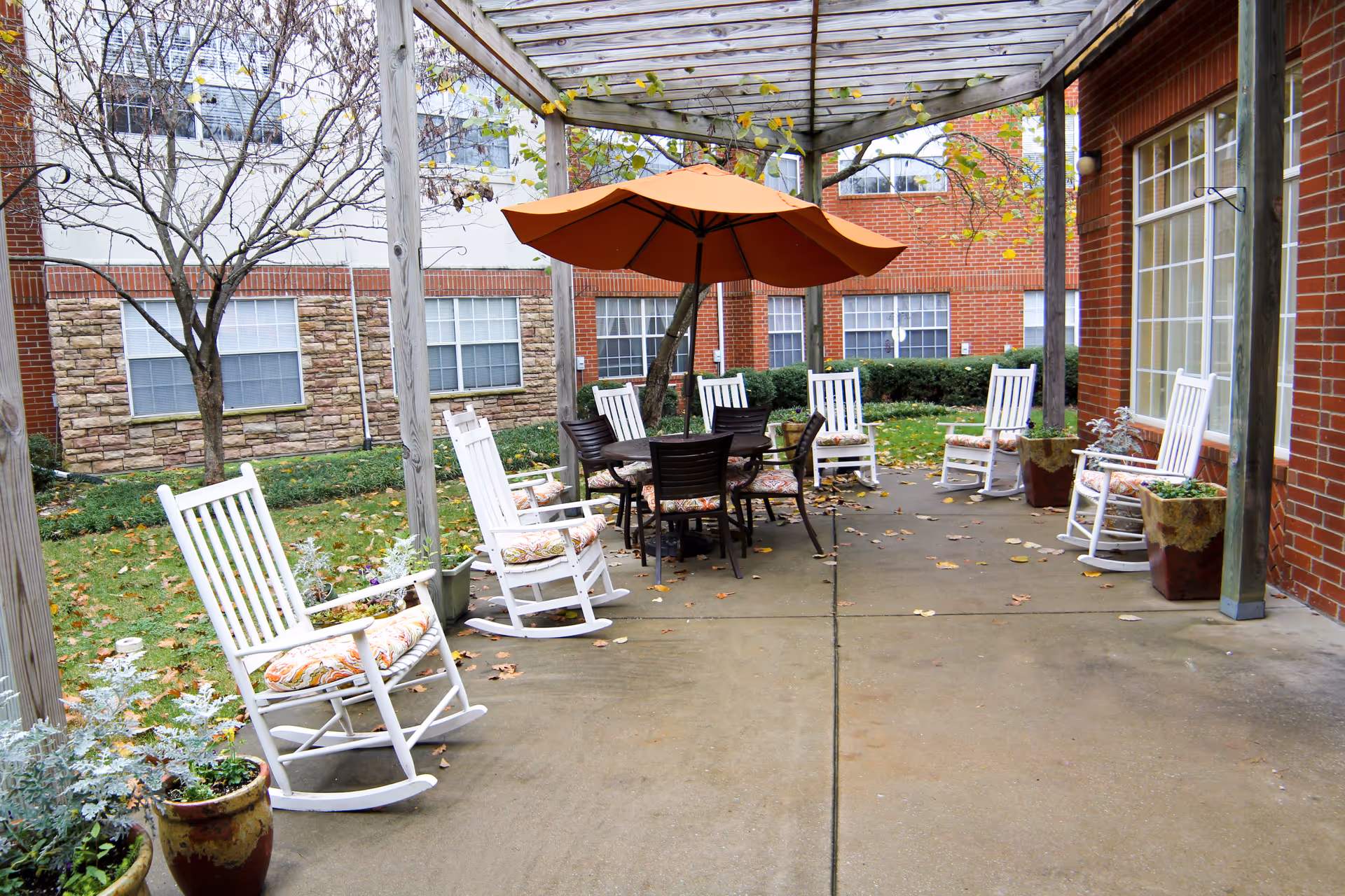 Outdoor courtyard patio with white rocking chairs around a central table under an orange umbrella.
