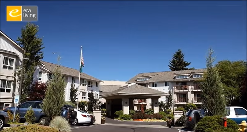 Front entrance of a multi-story senior living building with a covered porte-cochere, parked cars, landscaped beds and a flagpole under a clear blue sky.