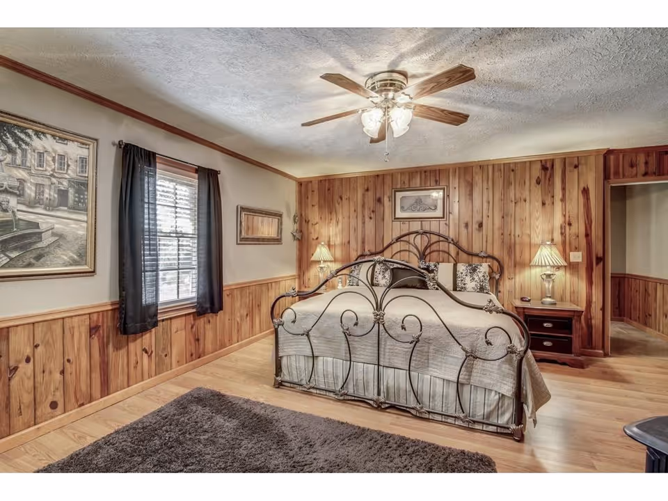 Rustic wood-paneled bedroom with an ornate iron bed, lamps, and a ceiling fan.