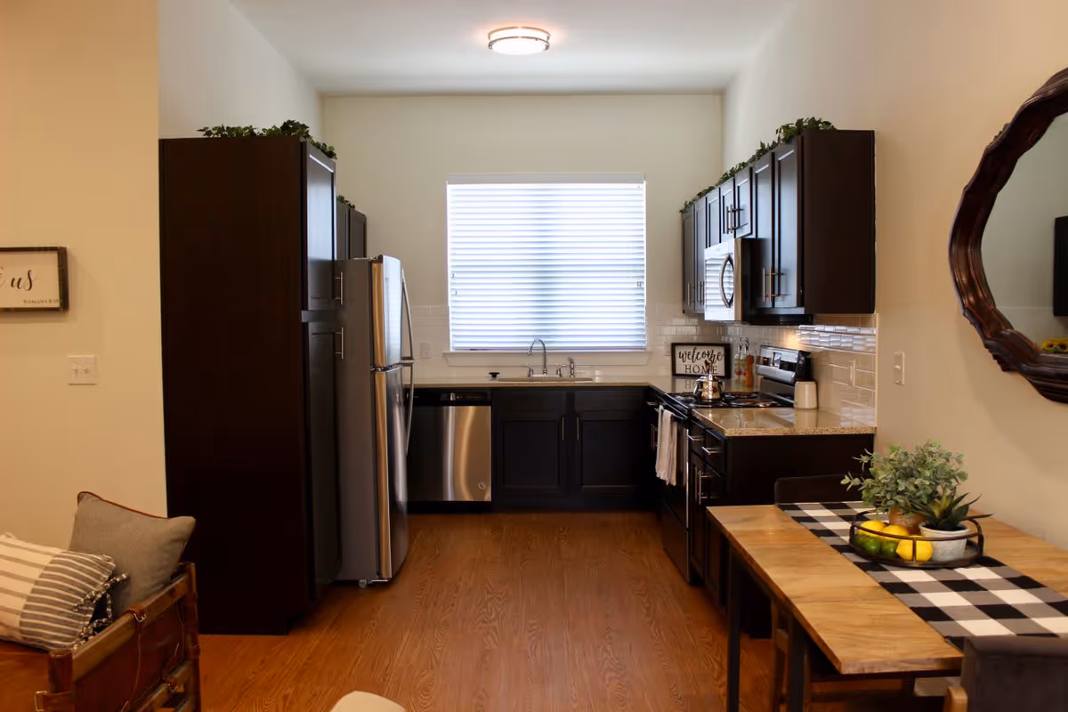 Open-plan kitchen with dark cabinets, stainless steel appliances, a sink under a window, and a small dining table.