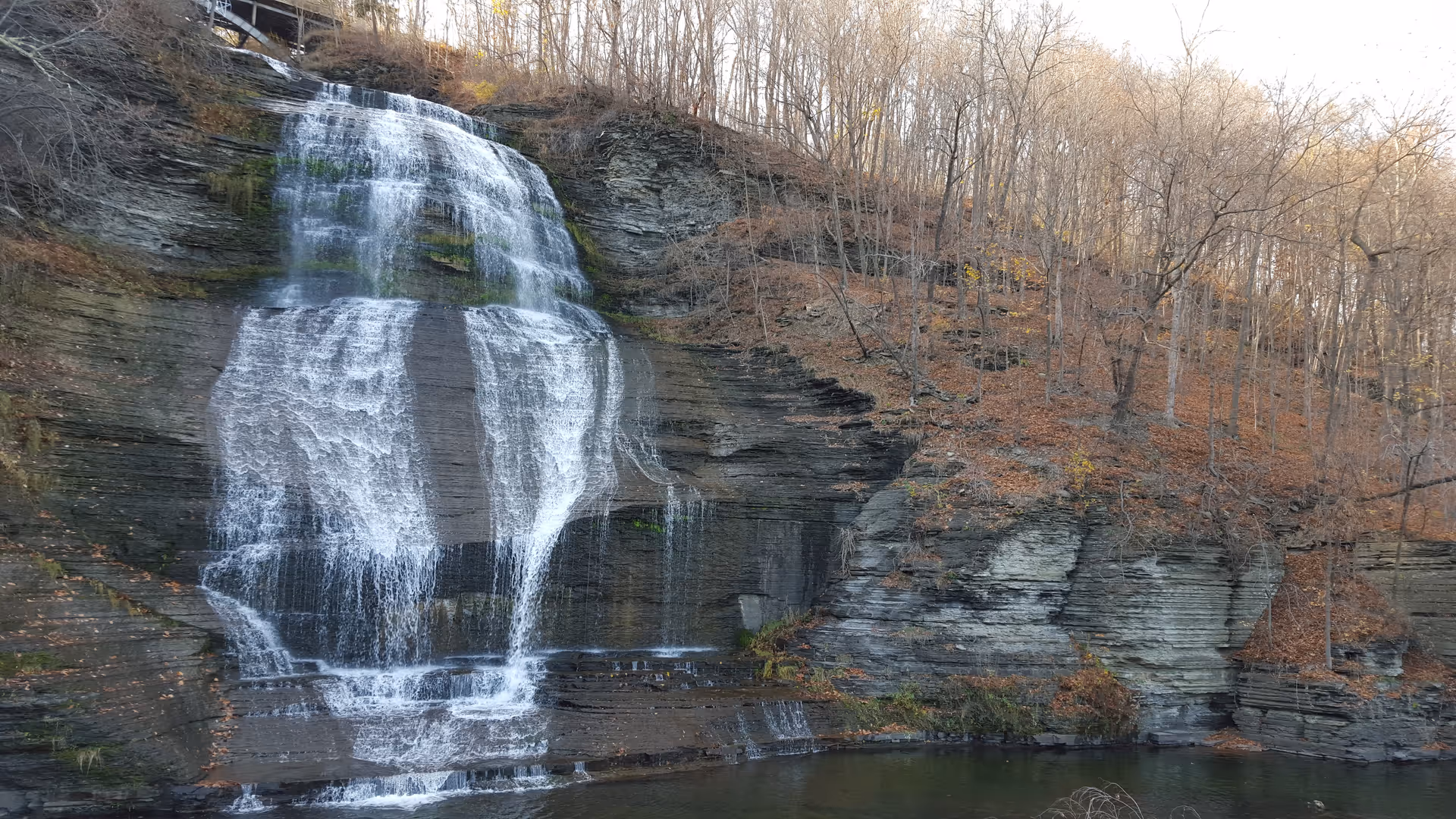 A waterfall spills over layered rocks beside leafless trees in a forested area.