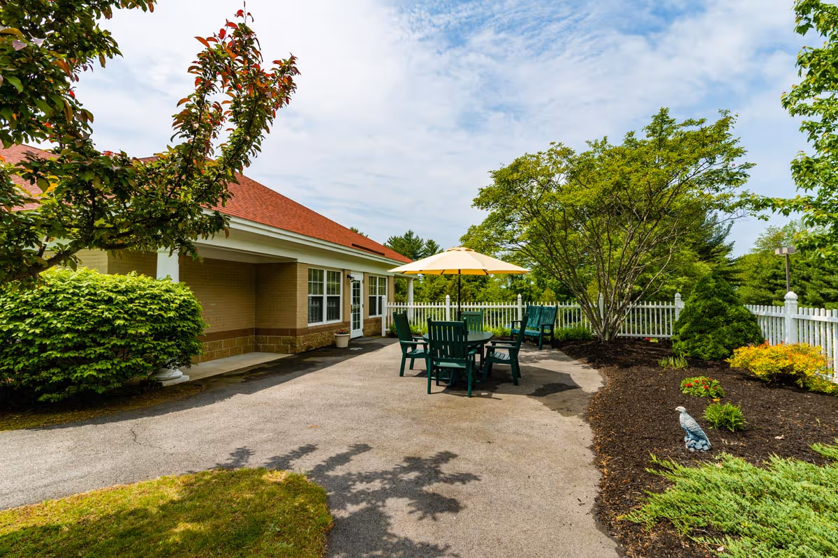 Outdoor patio area at St. Ann Rehabilitation and Nursing Center with green chairs and tables under a yellow umbrella, surrounded by trees, bushes, and a white picket fence.