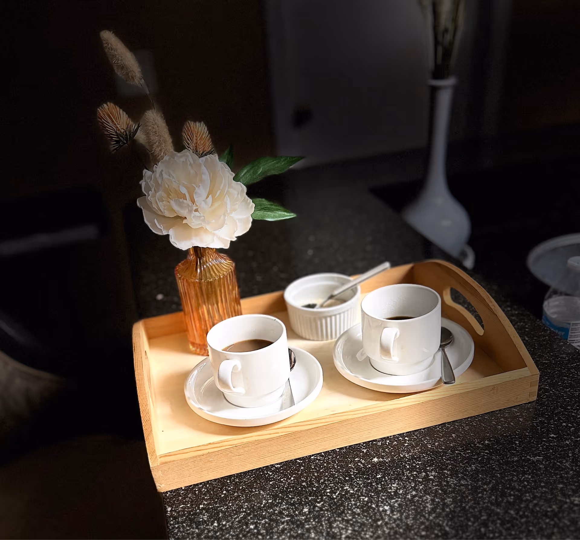 Wood serving tray with two coffee cups, saucers, spoons, and a flower vase on a counter