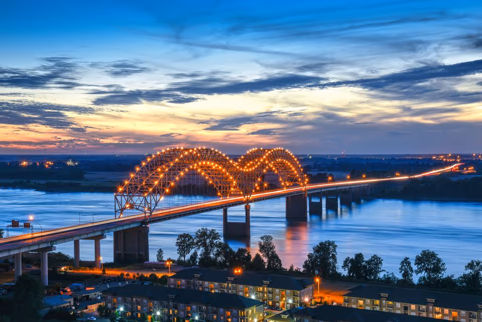 Sunset over a lit arched bridge spanning a wide river with buildings along the shore.