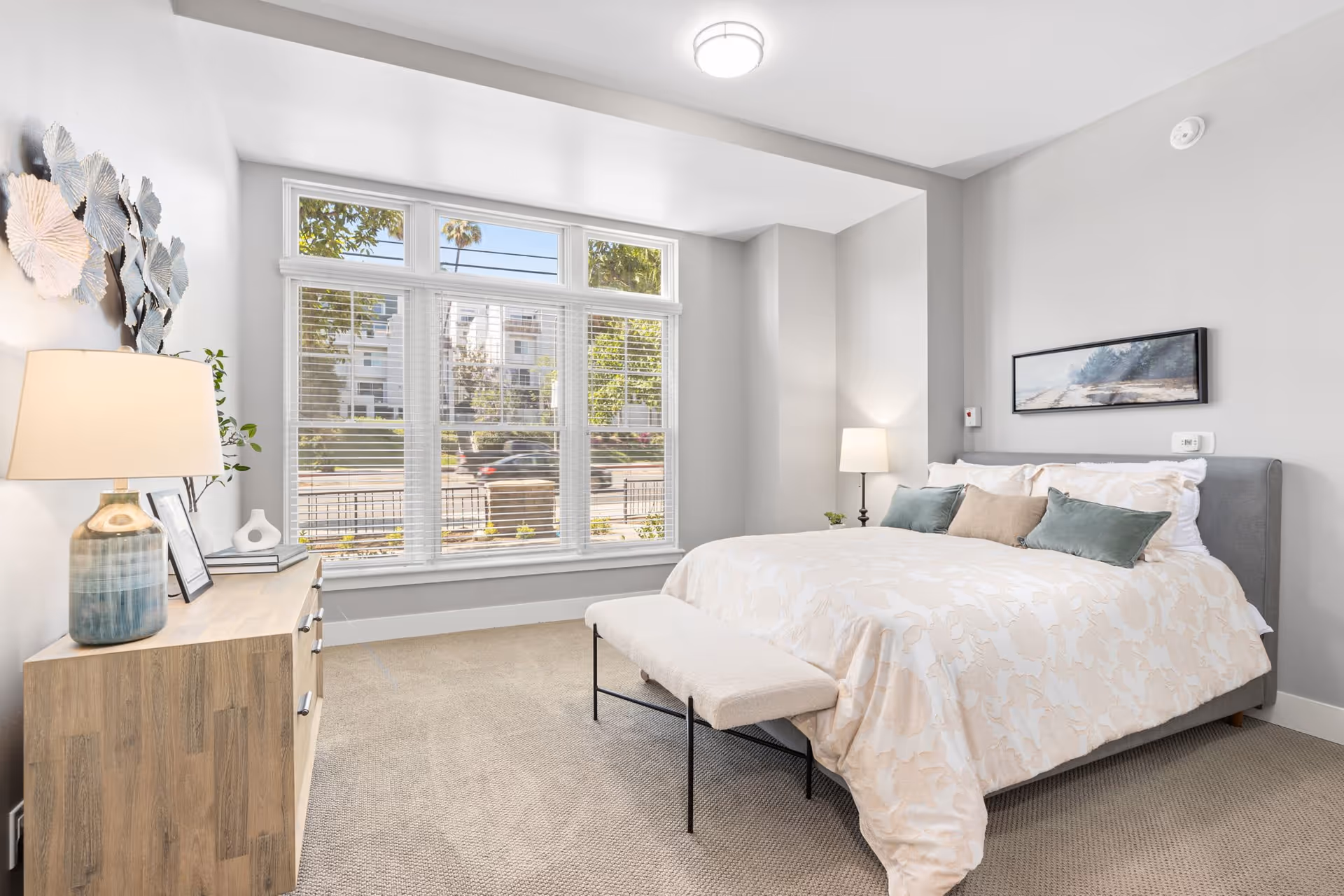 Bright and modern bedroom with a large window letting in natural light. The room features a neatly made bed with white and beige bedding, decorative pillows, a bench at the foot of the bed, a wooden dresser with a lamp and decorative items, and a wall-mounted artwork above the bed. The walls are painted light gray and the floor is carpeted.
