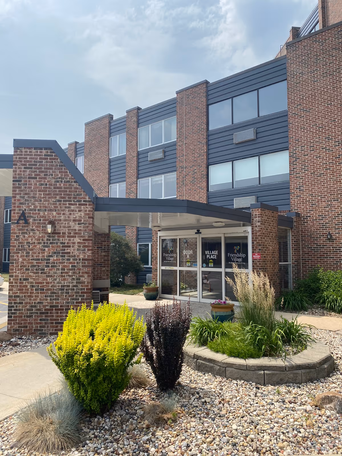 The entrance of a senior living facility featuring a brick facade, landscaped garden, and large glass doors.