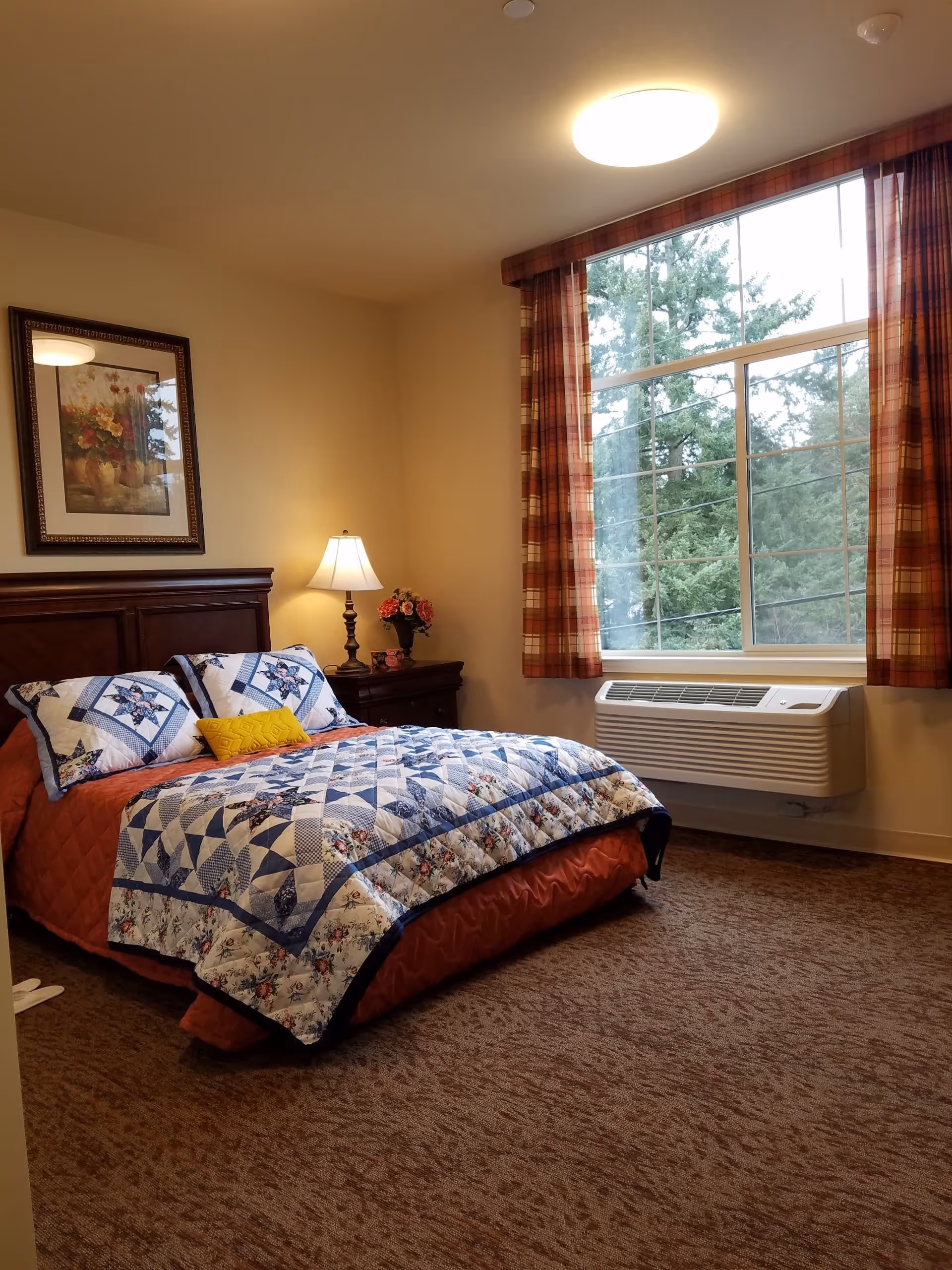 Sunlit bedroom with a made bed covered by a blue-and-white quilt, a bedside table with a lamp and flowers, and a large window with plaid curtains.