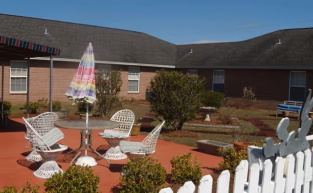 Outdoor patio area with white metal chairs and a round table with a colorful striped umbrella. The patio is surrounded by bushes and a white picket fence, with a brick building in the background under a clear blue sky.