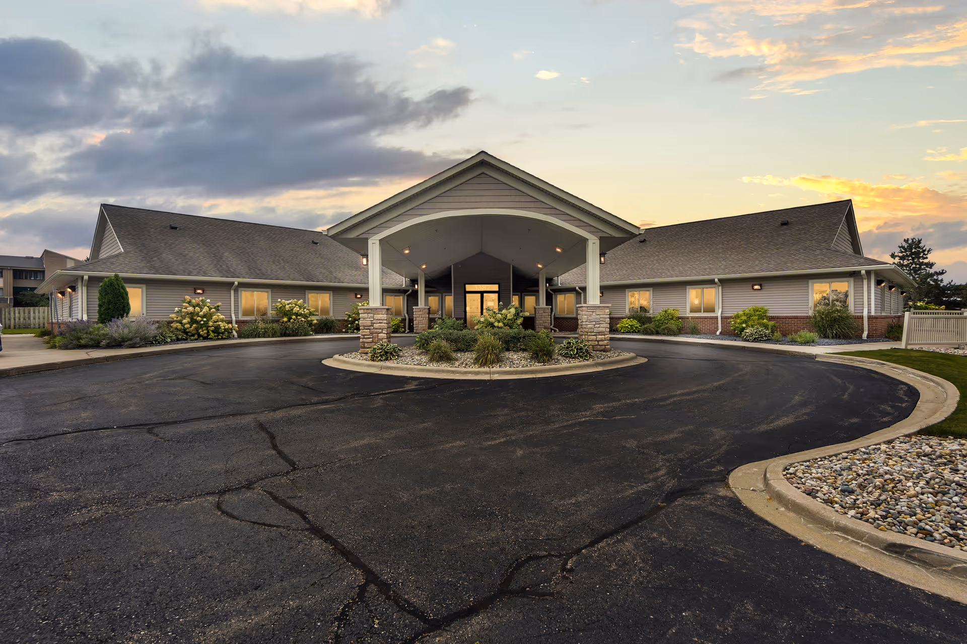 Single-story senior care facility with a covered porte-cochère entrance, circular driveway, and landscaped beds at sunset.