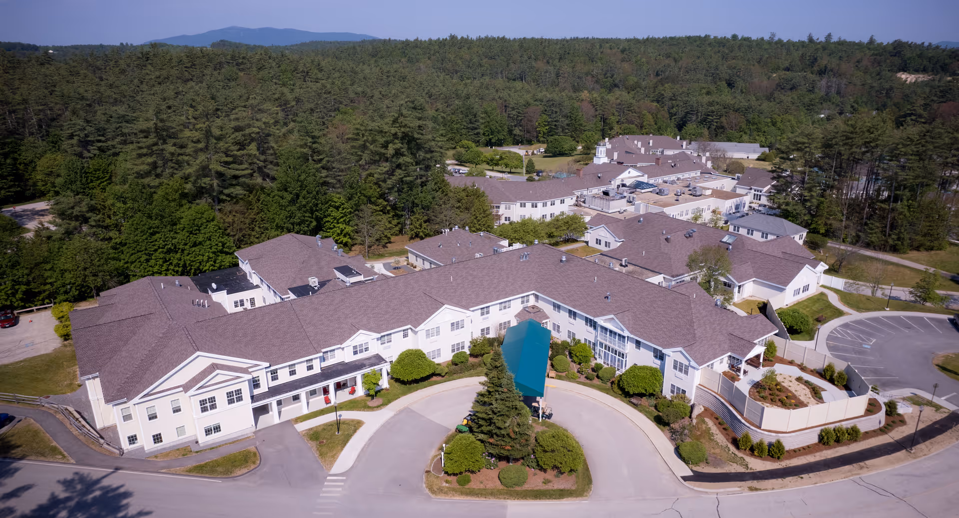 Aerial view of a large senior living facility with multiple connected buildings, a circular entrance drive, landscaped grounds, and surrounding woods.