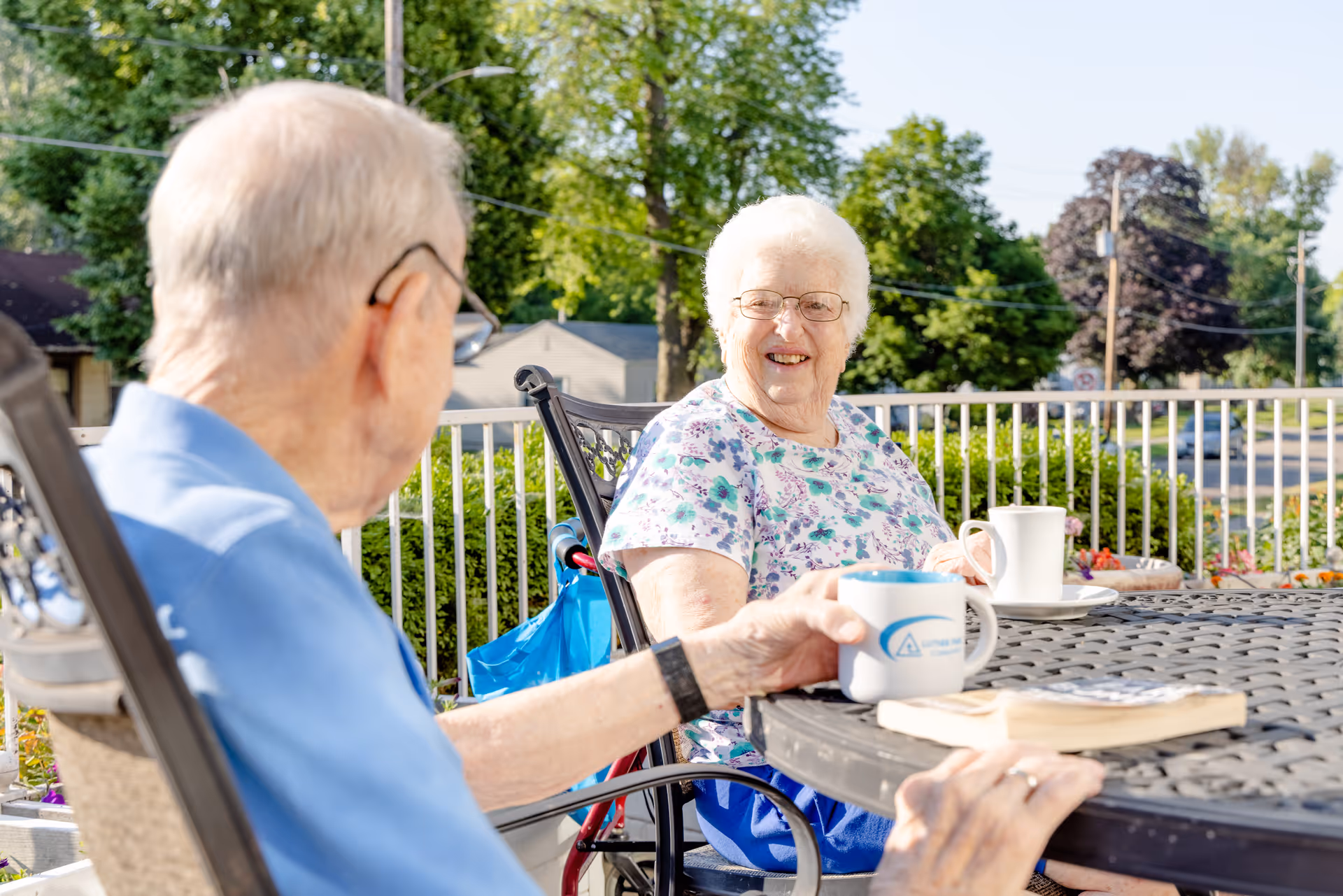Two elderly individuals sitting outdoors at a round metal table on a patio, enjoying drinks from mugs. One person is smiling and wearing glasses and a floral shirt, while the other person is seen from behind wearing a blue shirt. There are trees and bushes in the background under a clear sky.