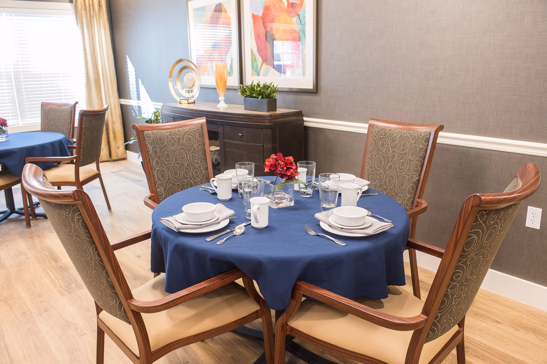 Round dining table with a blue tablecloth set for four, surrounded by upholstered wooden chairs in a well-lit dining room with artwork and a sideboard.