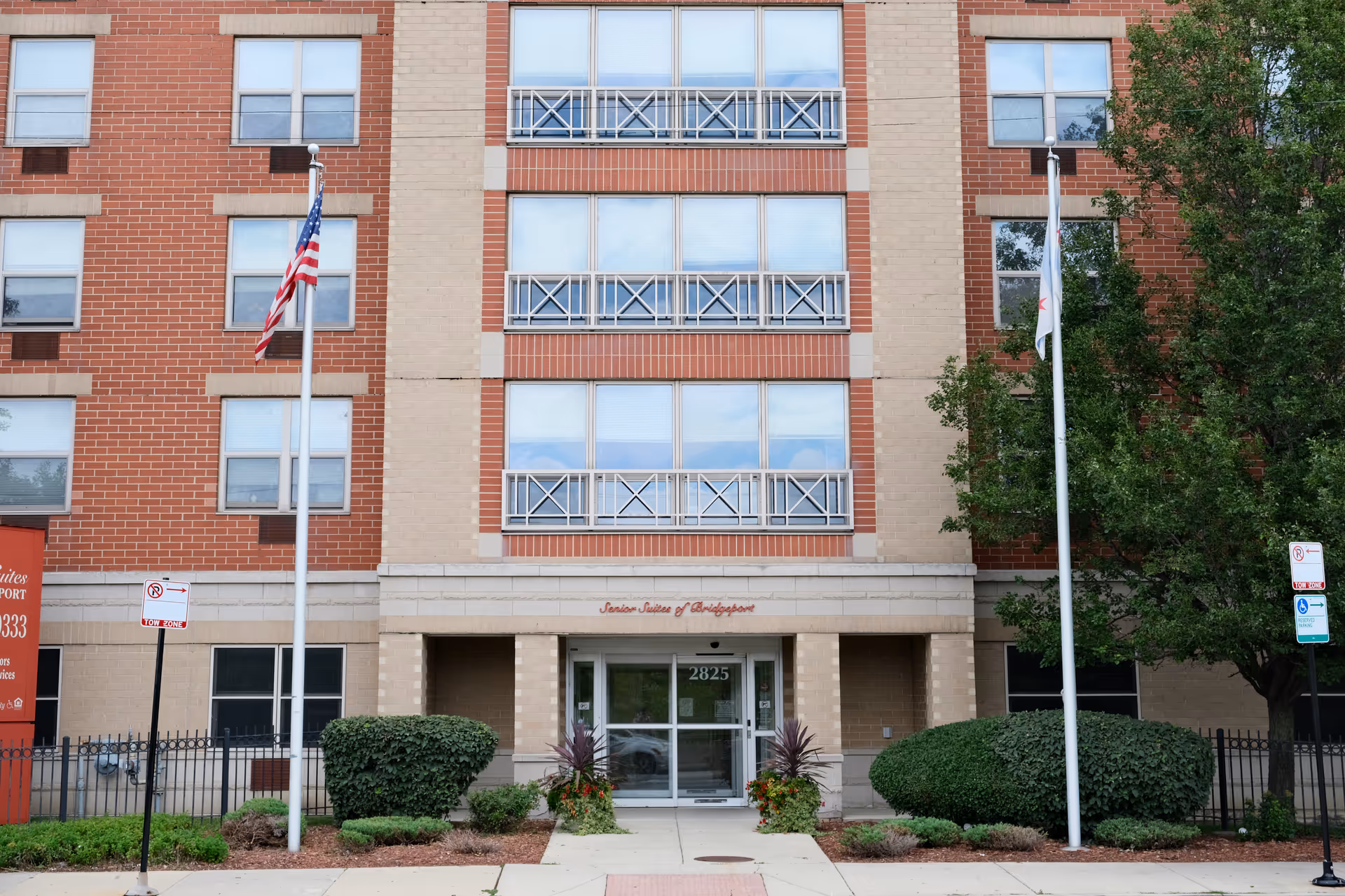Front exterior view of Senior Suites of Bridgeport, a multi-story brick and beige building with large windows and balconies. Two flagpoles with flags stand on either side of the entrance, which has glass doors and the address number 2825 above it. There are trimmed bushes and small plants along the walkway leading to the entrance.