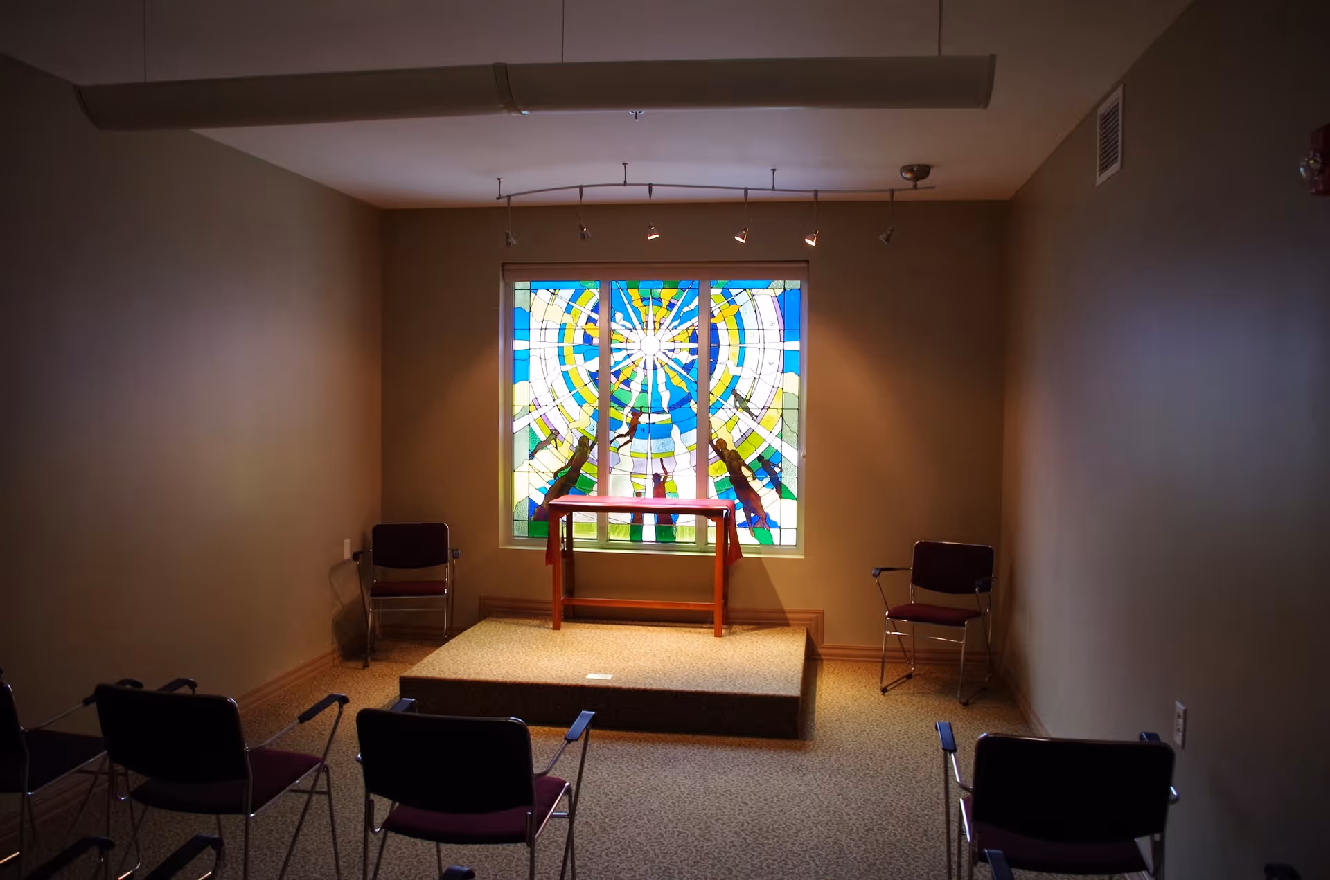 Small chapel-style room with a colorful stained-glass window, a raised platform and table, and chairs arranged facing it.