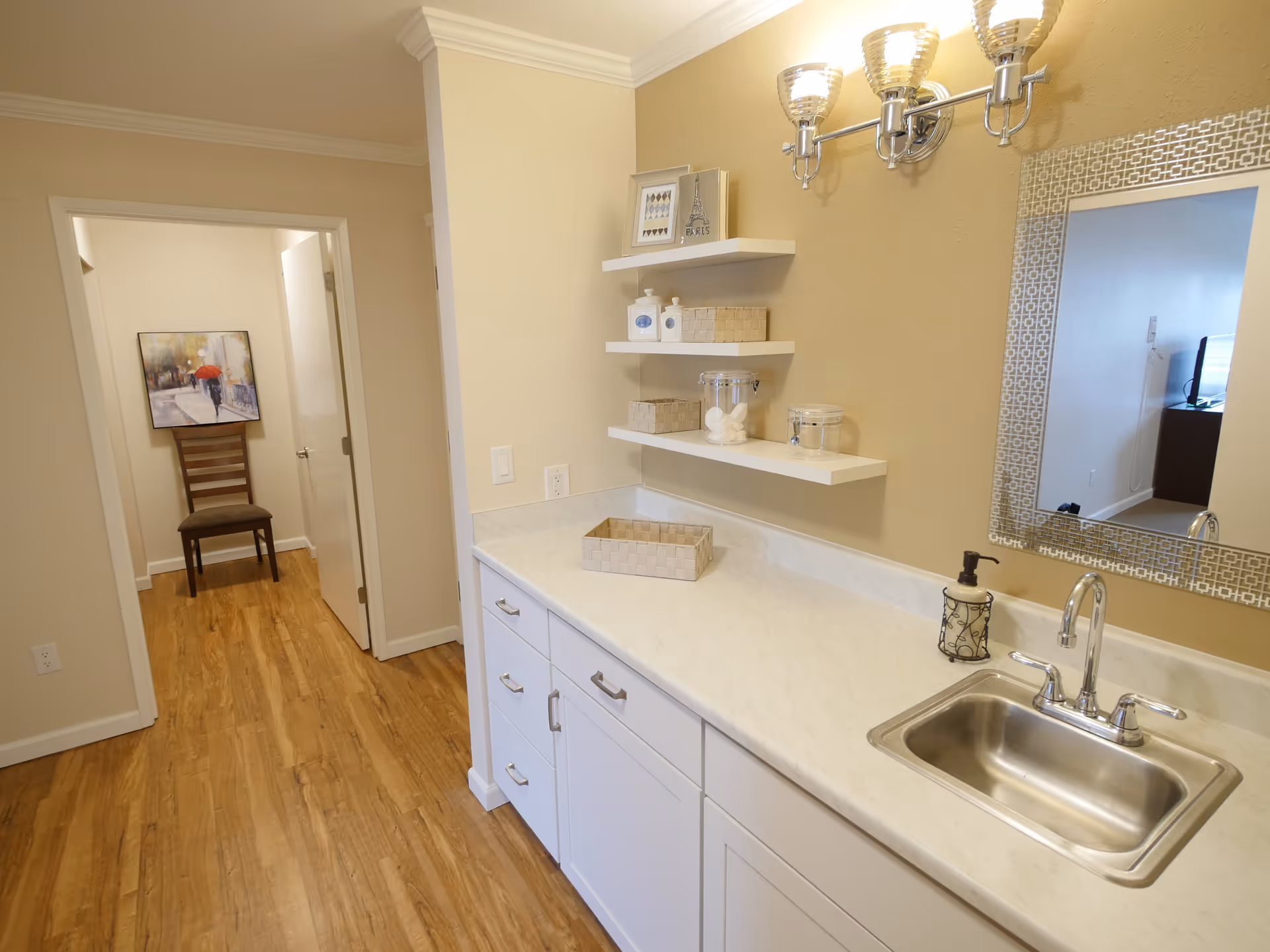 Interior view of a room with a white countertop and sink on the right side, three floating shelves with decorative items above the counter, a large mirror on the wall, and a hallway leading to another room with a chair and a painting of a person with a red umbrella.