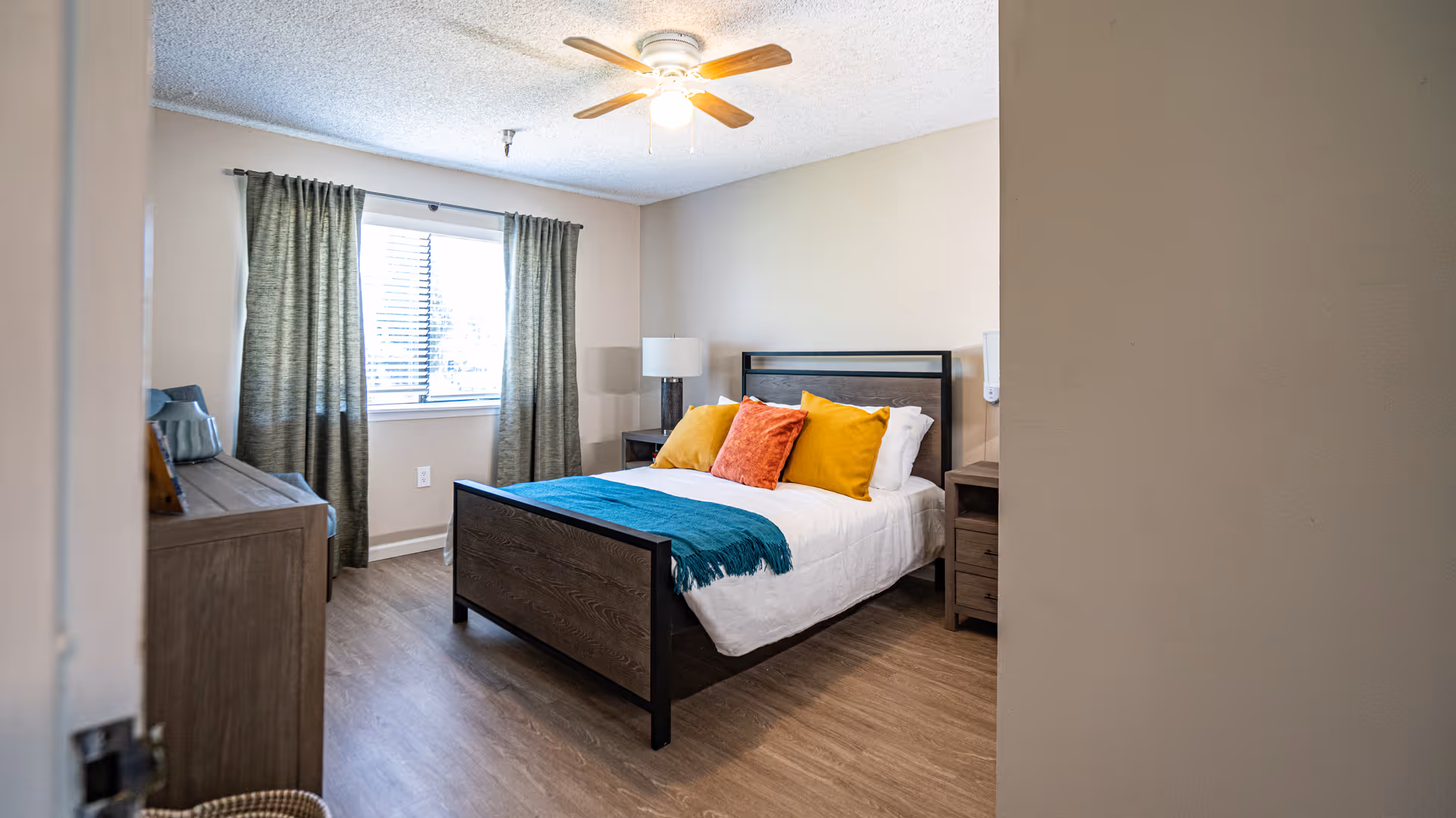 Well-lit furnished bedroom with a bed topped by colorful pillows, a dresser, window with curtains, and a ceiling fan.