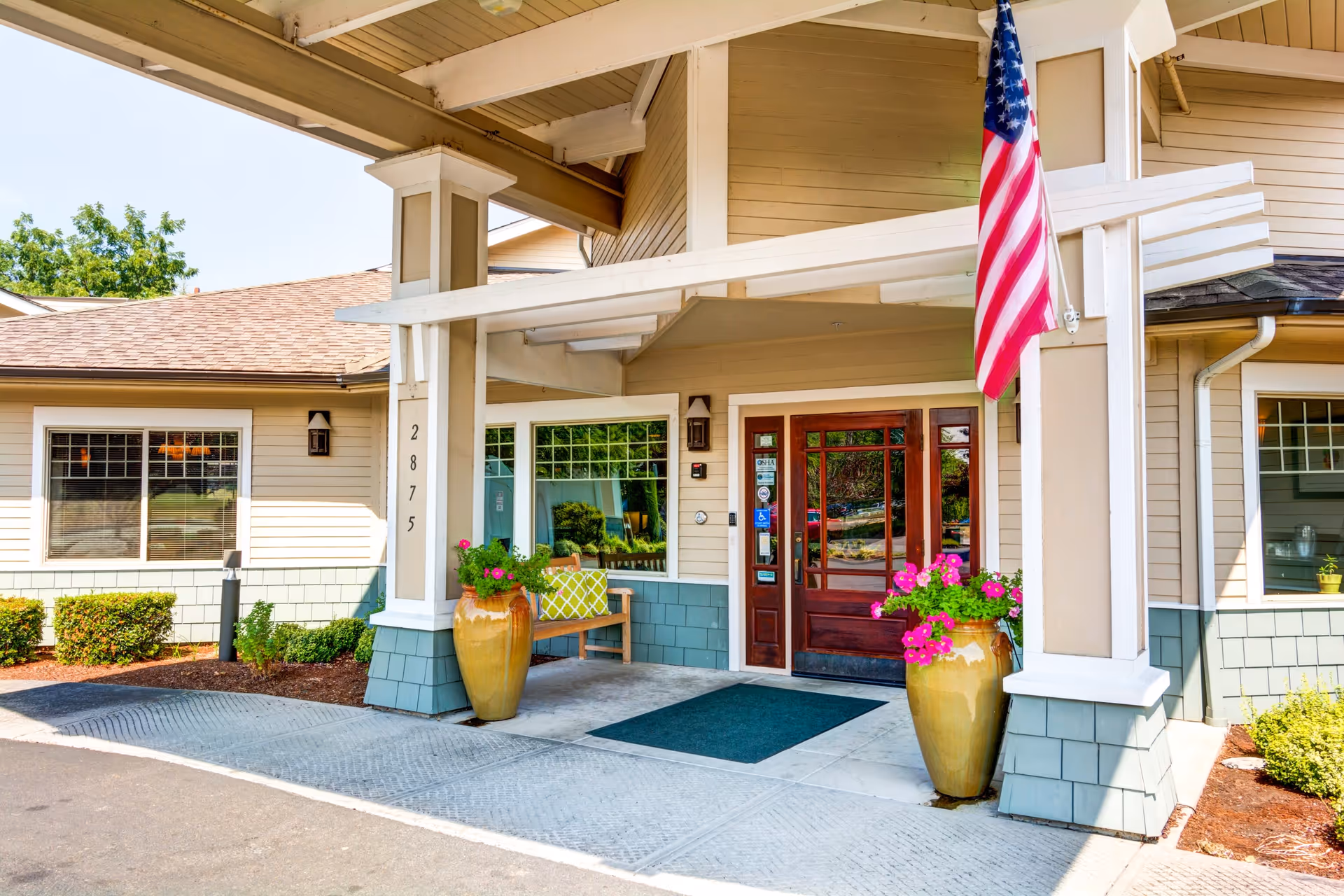 Entrance of Timberwood Court Memory Care facility featuring a covered porch with two large flower pots containing pink flowers, a wooden bench with a green and white cushion, an American flag mounted on a pillar, and a glass door with wooden framing. The building exterior has beige siding with green shingle accents and several windows.