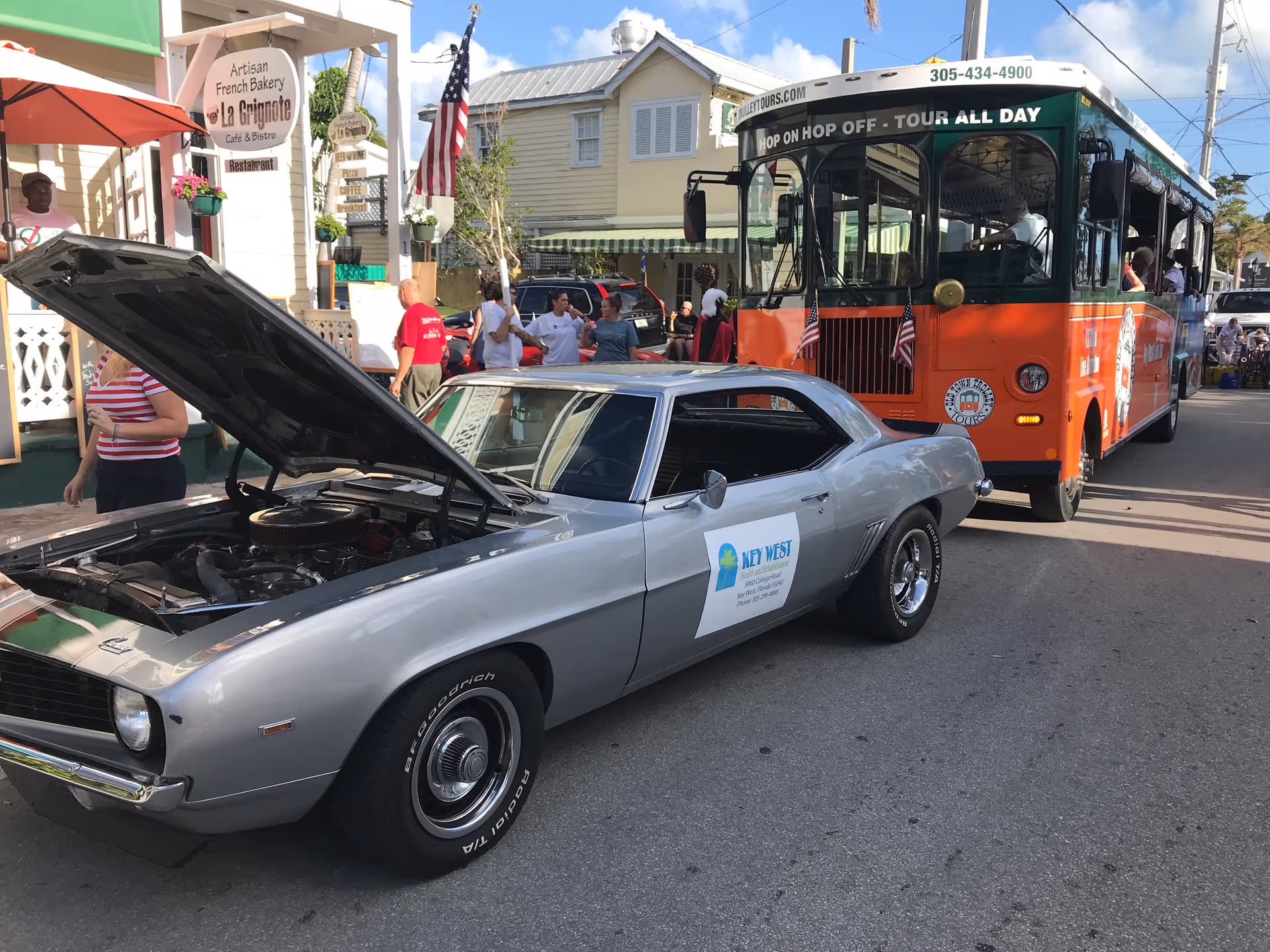 A silver vintage car with hood open beside an orange tour bus on a sunny street.