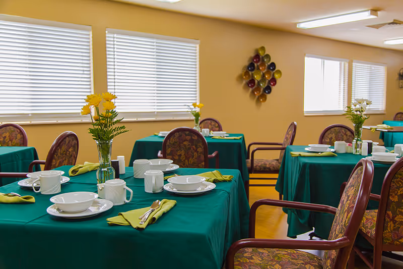 A dining room with multiple tables covered in green tablecloths, each set with white plates, bowls, cups, and silverware with green napkins. There are floral centerpieces on each table, and the chairs have patterned upholstery. The room has beige walls and windows with white blinds.