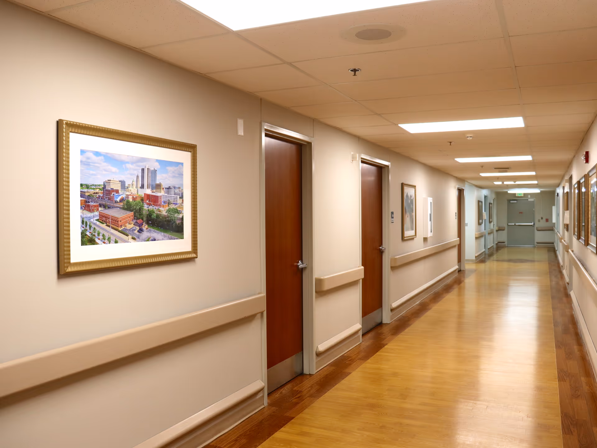 A long, well-lit hallway in a senior living facility with wooden doors on both sides, beige walls with handrails, framed pictures hanging on the walls, and a polished wooden floor.