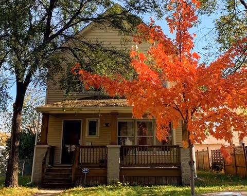 Two story beige house with a front porch and orange fall foliage in the yard.