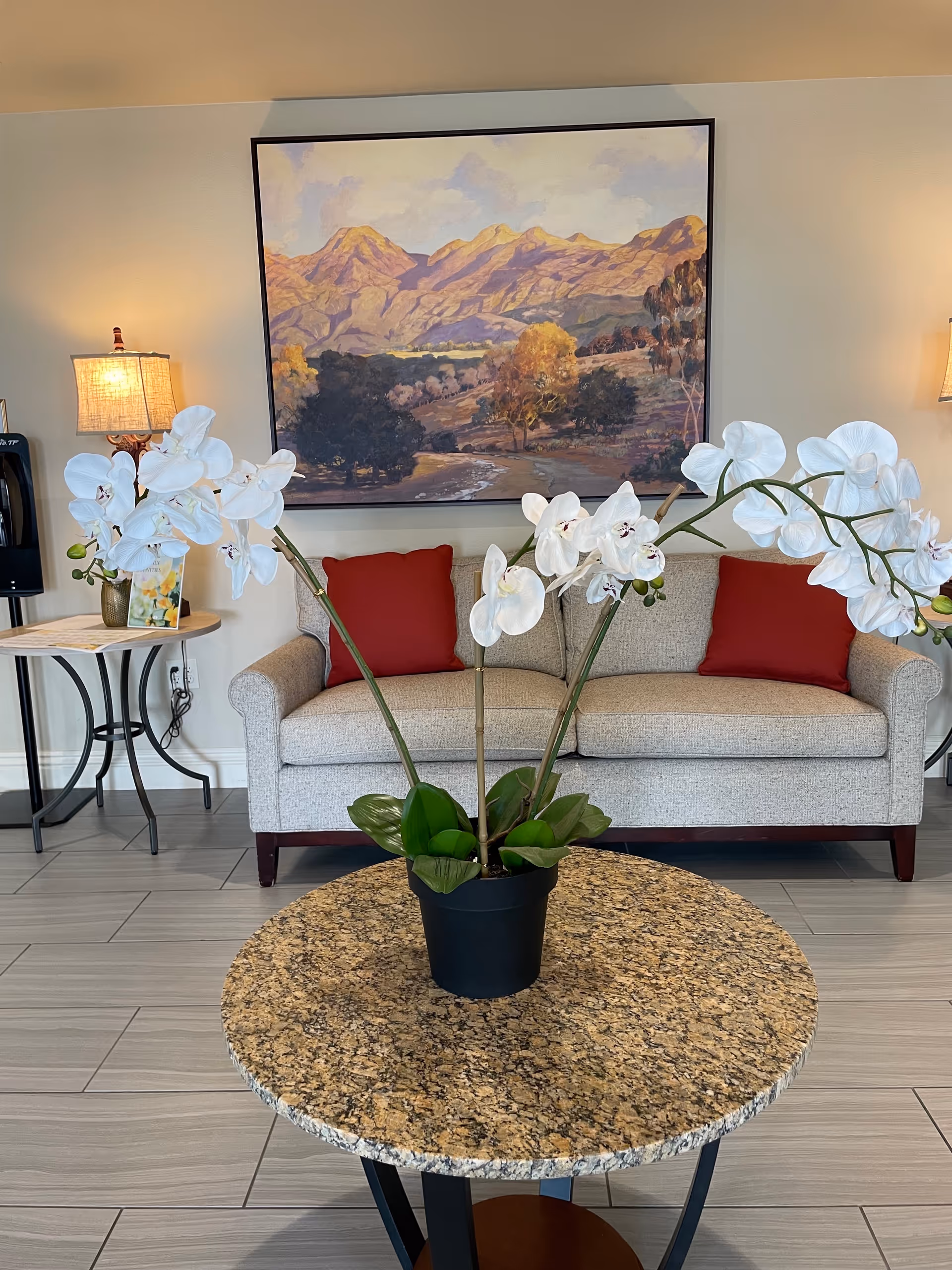 Lobby seating area with a gray sofa, red cushions, mountain painting, and white orchids.