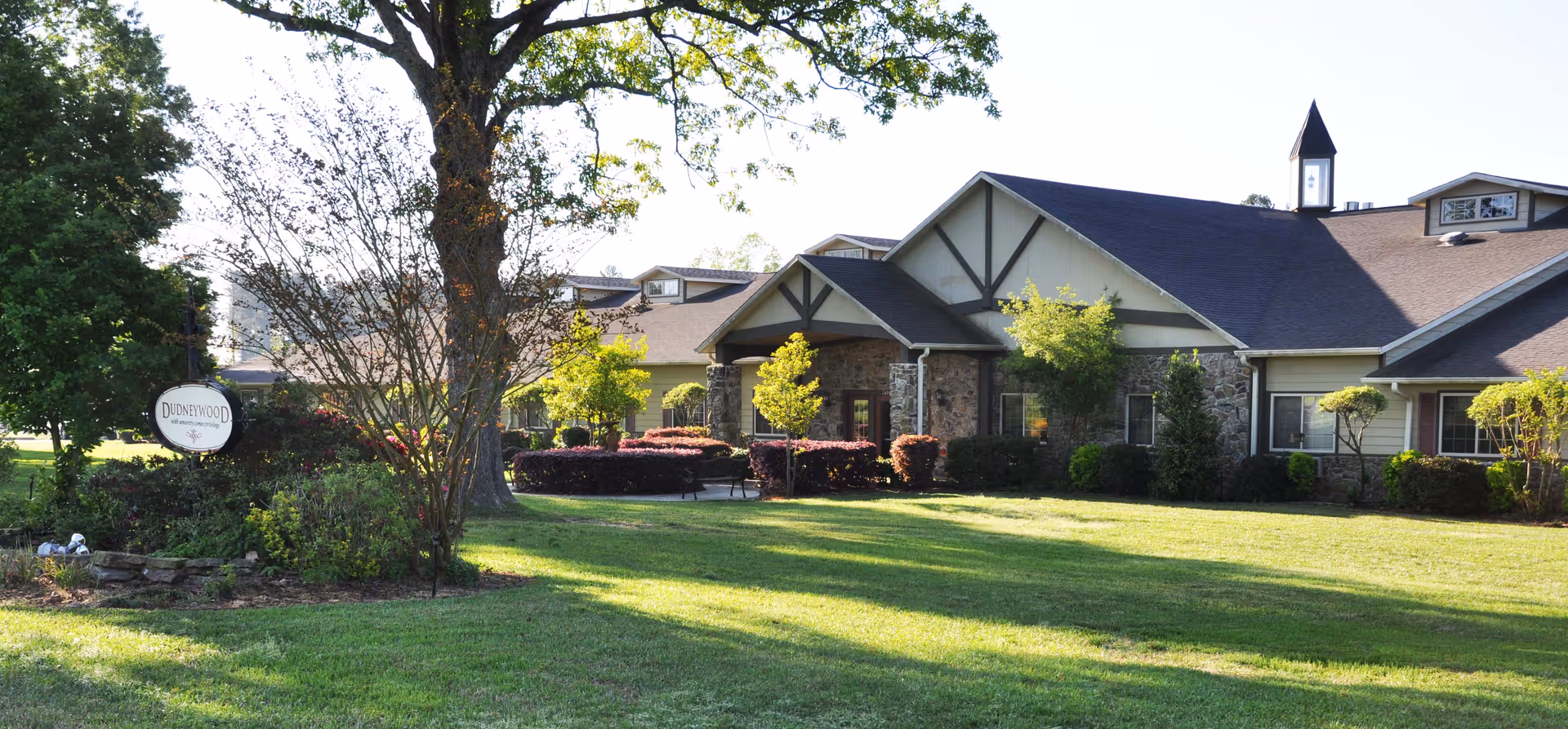 Exterior view of Dudneywood Assisted Living facility showing a single-story building with stone and siding facade, a peaked roof with a small cupola, surrounded by well-maintained green lawn, trees, and shrubs under a clear sky.