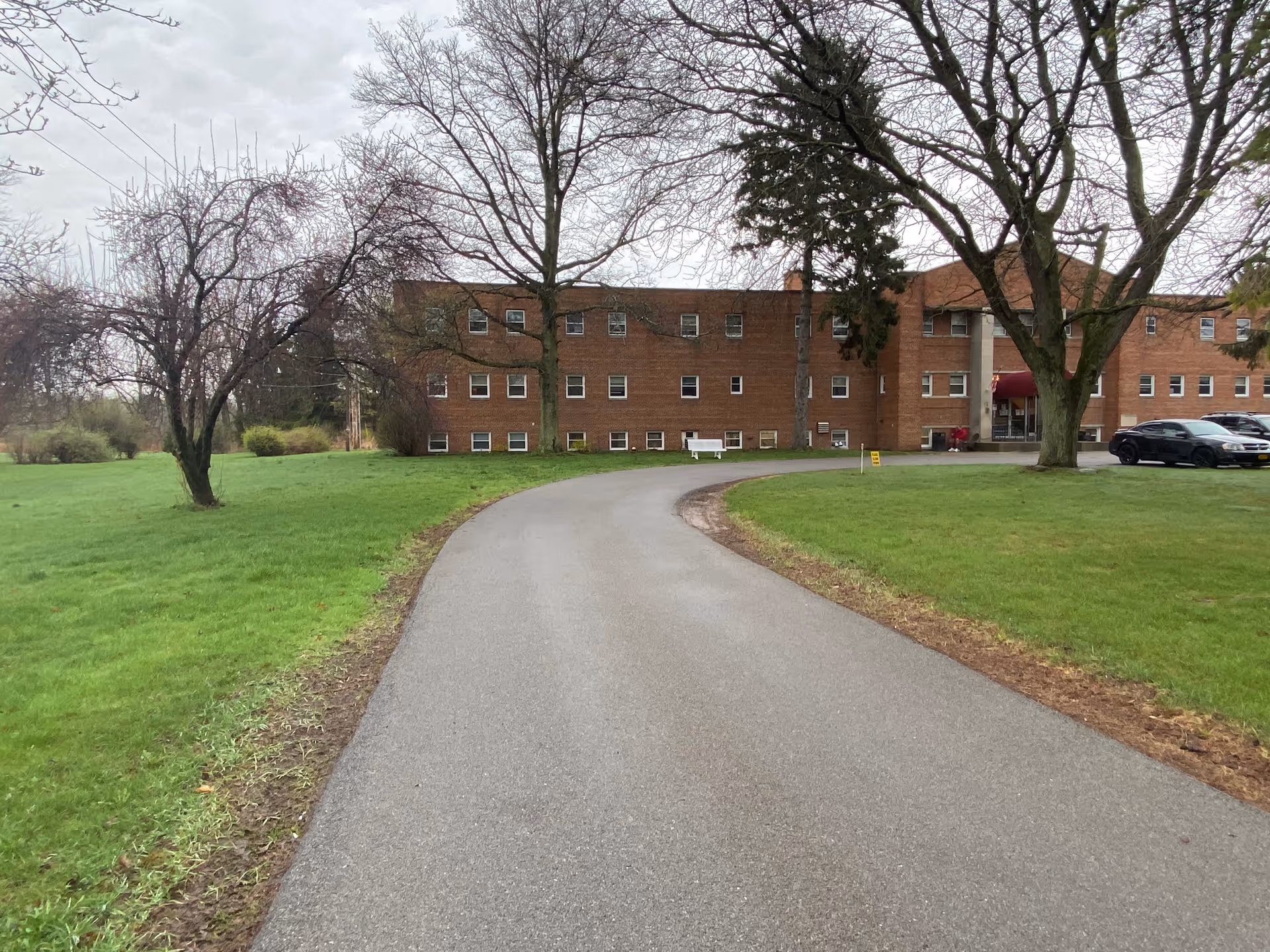 Front view of Leroy Manor, a brick senior living building, with curved driveway, lawn, and trees.