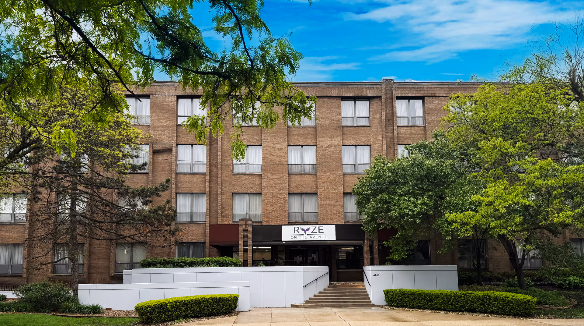 Exterior view of Ryze on the Avenue senior living building with trees and entrance steps