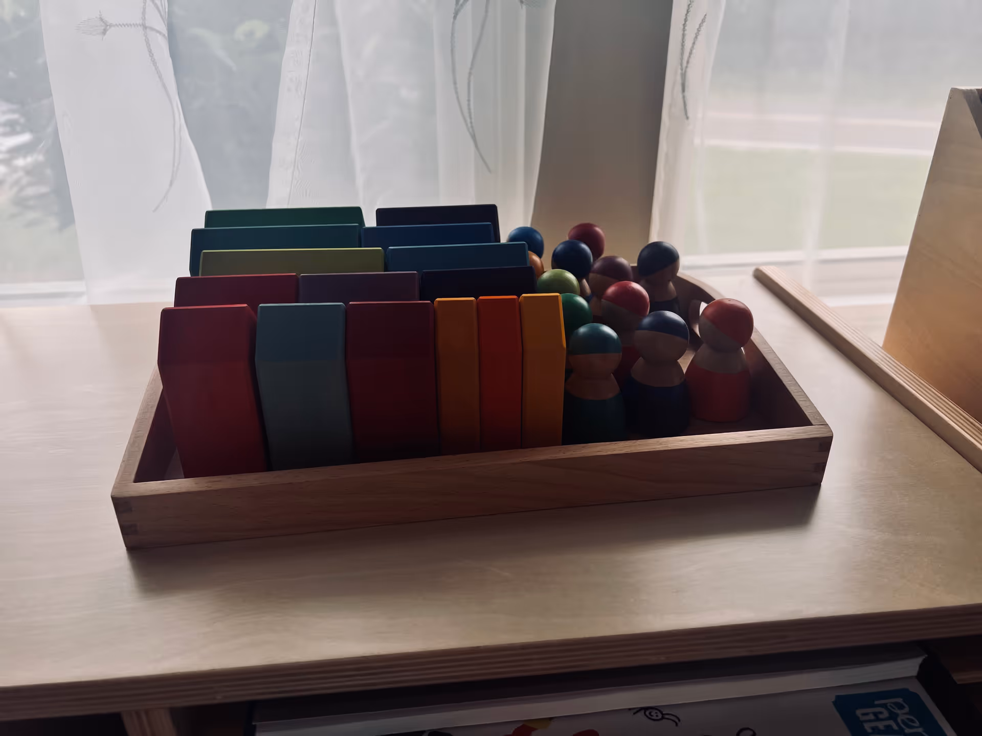 Wooden tray with colorful blocks and peg figures on a table by a window.