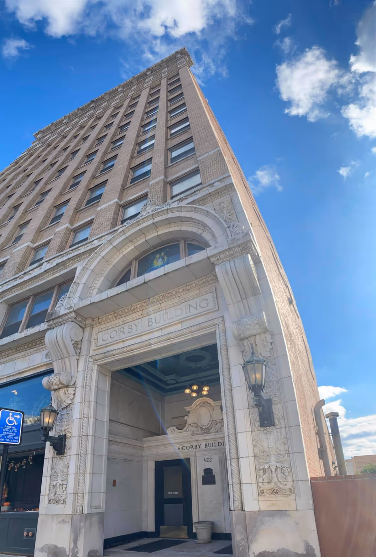 Upward view of the Corby Building, a tall multi-story brick building with ornate white stone detailing around the entrance, under a blue sky with some clouds.