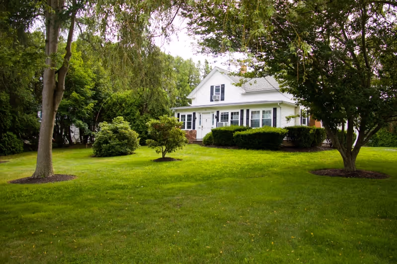 A white house with black shutters surrounded by a well-maintained green lawn and several trees and bushes under a cloudy sky.