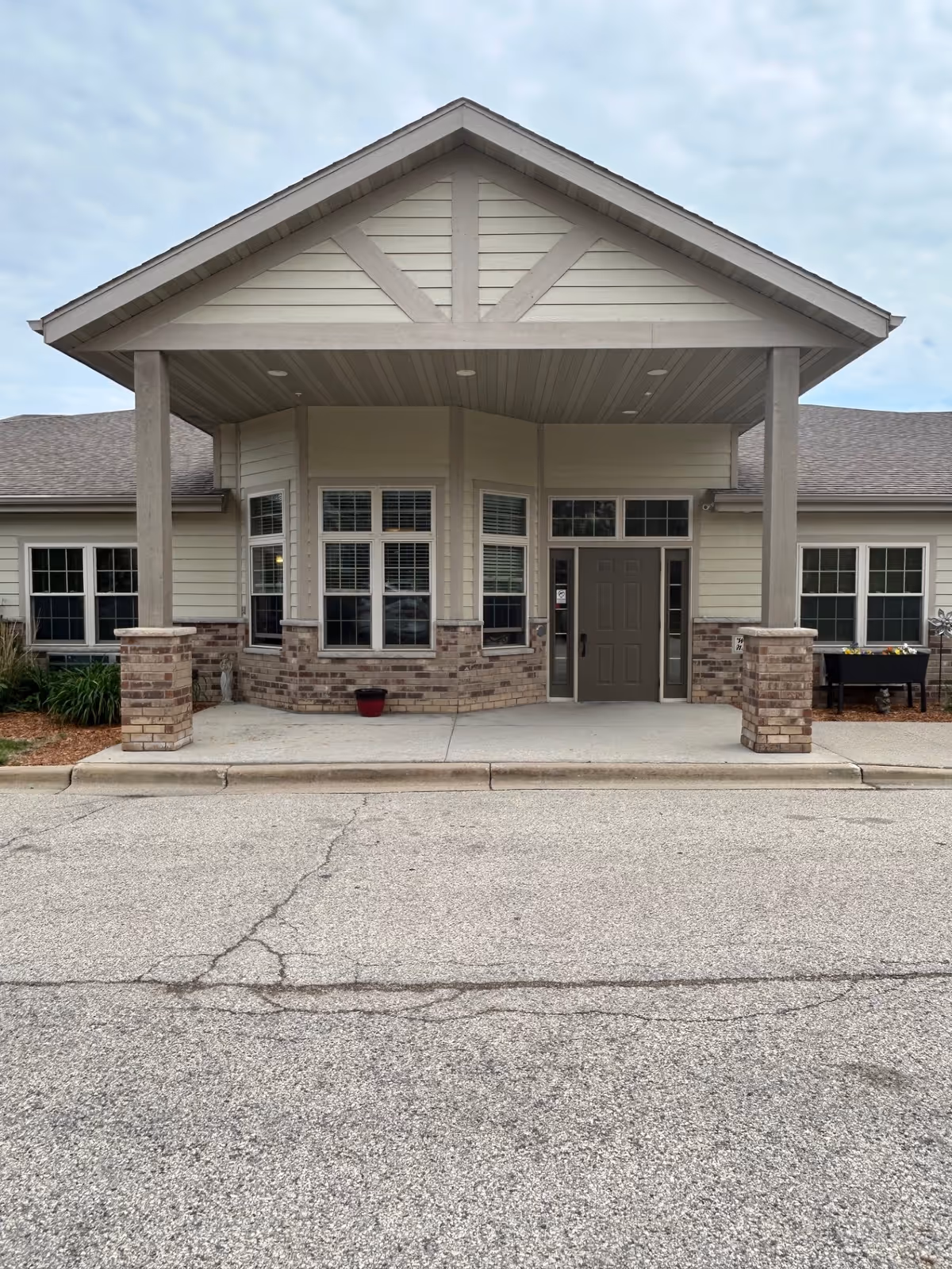 Front entrance of a single-story building with a covered porch supported by four columns. The building has beige siding with brick accents around the lower walls and multiple windows with white frames. The entrance door is centered under the porch roof, and there is a paved driveway or parking area in front.