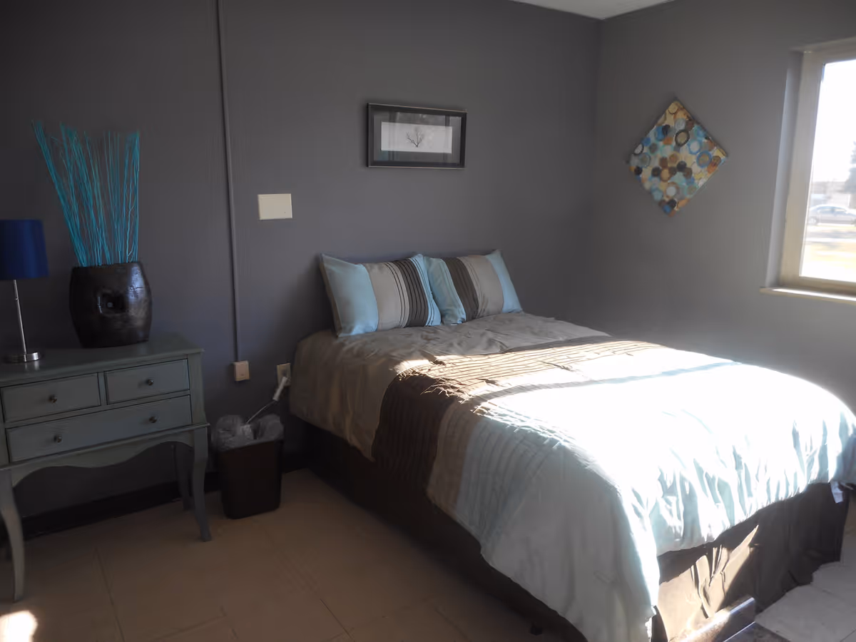 A sunlit bedroom with a made bed, bedside table with a lamp and decorative vase, and wall art on gray walls.
