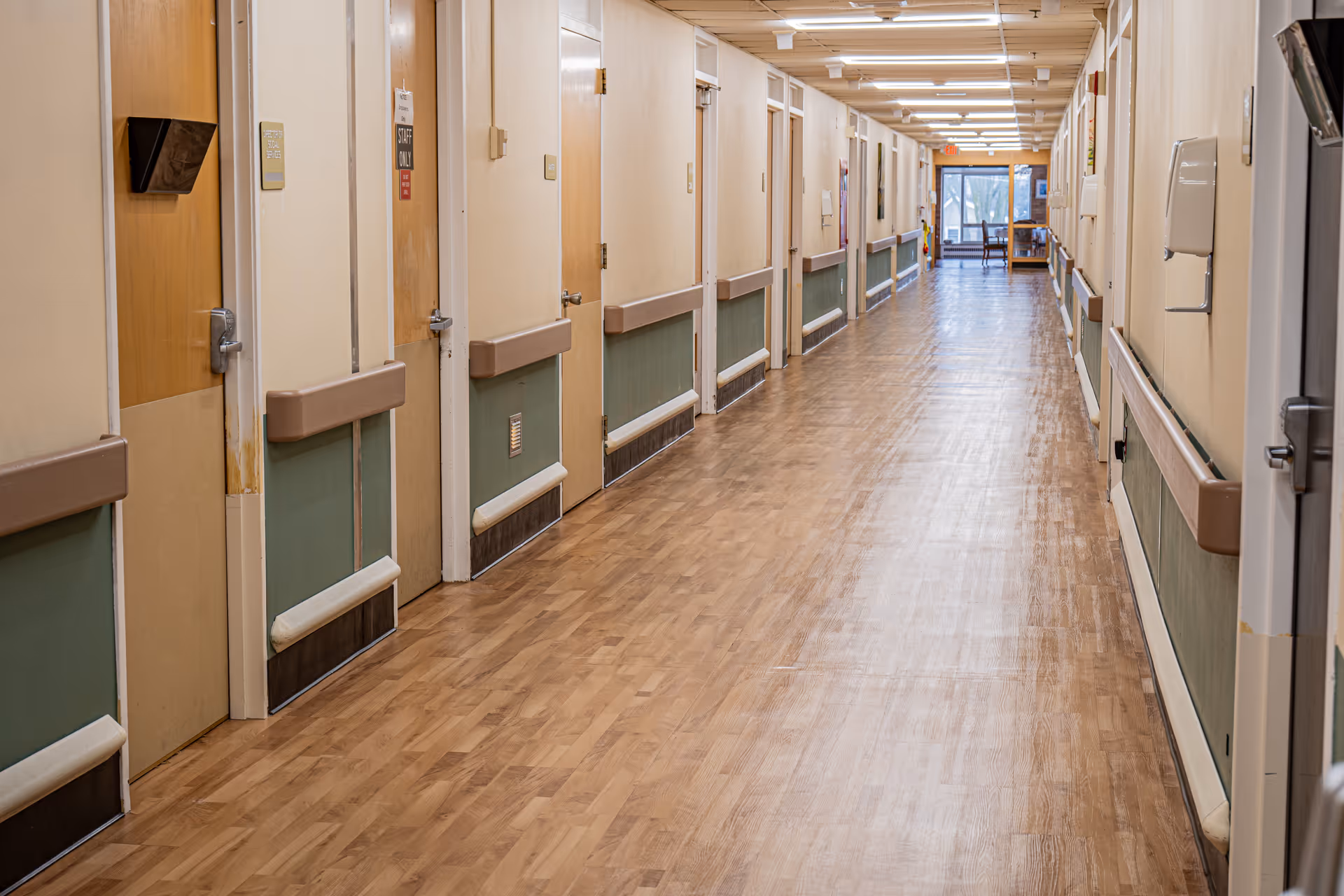 A long, empty hallway in a senior living facility with multiple closed doors on both sides. The floor is wooden, and the walls have handrails for support. At the end of the hallway, there is a window with natural light coming through and a small seating area with chairs and a table.