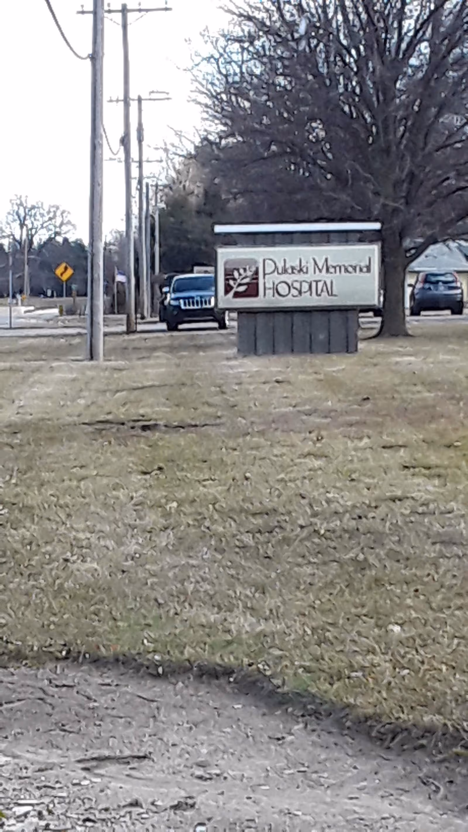 Outdoor sign for Pulaski Memorial Hospital beside road with leafless trees and cars.